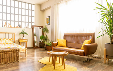 Interior home design. Spacious and bright boho-style bedroom, in an industrial loft, with wooden furniture: bed, night tables, brown sofa, yellow carpets, large windows and indoor green plants.
