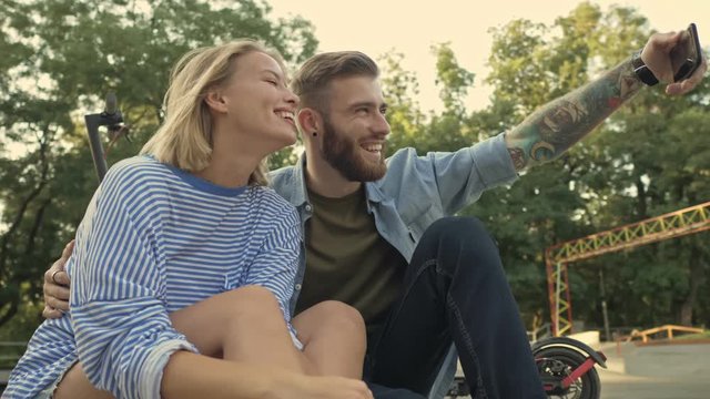 A young happy couple is taking a selfie while resting after driving electric kick scooters on the sport area