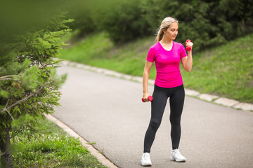 beautiful young woman in sports pink t-shirt   with dumbbells in hands in the summer park