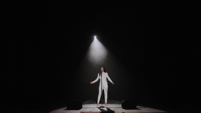 A beautiful singer with long hair sings on stage in the dark under the light of a white spotlight. Girl in a white suit performing on stage. Female singer on the stage holding a microphone.