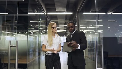 Good-looking serious 30s multiethnic business people standing in glass office hall and discussing aboult business project using tablet pc