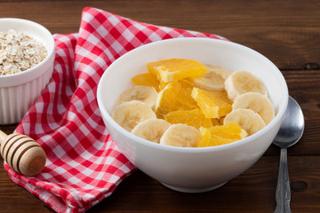 Bowl of oatmeal porridge with banana and orange  on wooden background, copy space