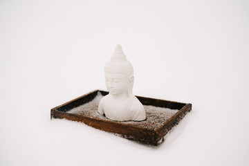 a small statue of a white Buddha made of plaster on a wooden stand covered with white snow during a...