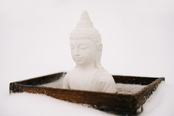 a small statue of a white Buddha made of plaster on a wooden stand covered with white snow during a snowfall in winter