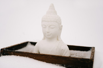 a small statue of a white Buddha made of plaster on a wooden stand covered with white snow during a...