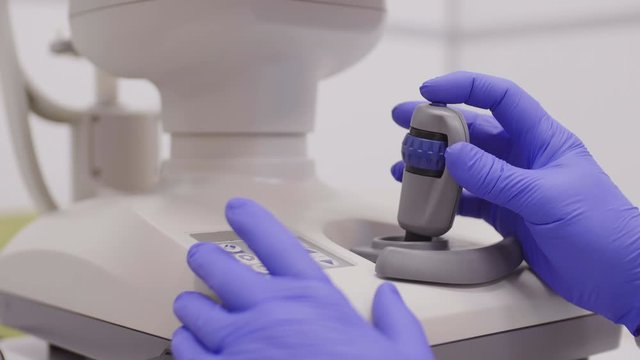 Close up shot of hands of optometrist in disposable gloves using joystick lever of autorefractor machine while giving eye examination in clinic