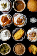 Homemade citrus oranges or clementines sweet muffins cupcakes in baked tray with different cream, pistachio, caramel toppings above over wooden cutting board as background. Flat lay, close up