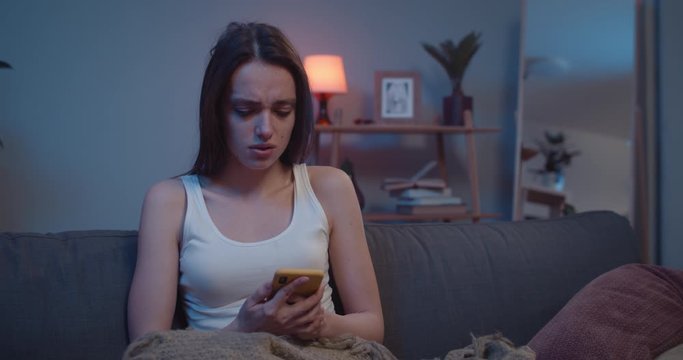 Unhappy young girl getting bed news on her smartphone while sitting on sofa at home . Female millennial looking worried and sad while checking mobile phone content. Indoors.