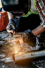 Close up portrait view of professional mask protected welder man in uniform working on the metal sculpture at the table in the industrial fabric workshop in front of few other workers.