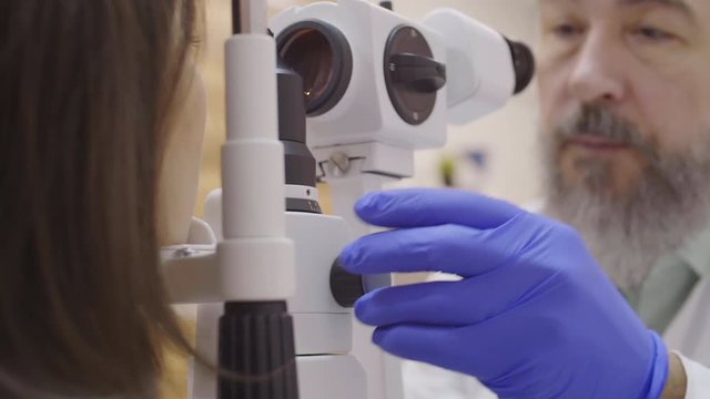 Over the shoulder shot of elderly male optometrist in disposable gloves looking through oculars of slit lamp while examining retina of female patient