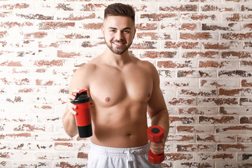 Sporty man with protein shake and dumbbell on brick background