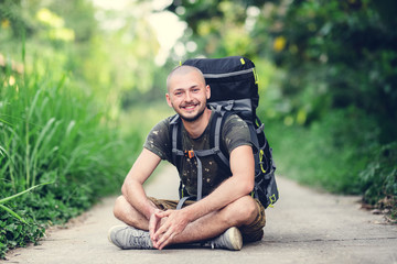 Young white Caucasian man with a large hiking backpack behind his back smiling while sitting on a jungle trail. The traveler smiles.