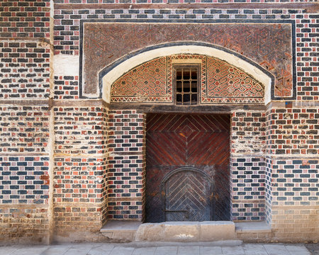 Grunge wooden decorated arched entrance gate with inner small wooden door on wall with black and red bricks with white seam
