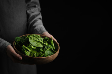 wooden plate with fresh spinach leaves in the hands of a woman dressed in gray linen clothes....