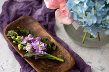 Purple Flowers in Small Wooden Tray on White Table; Purple Fabric in Background; Blue Hydrangea and Pink Roses in Bouquet in Background