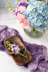 Purple Flowers in Small Wooden Tray on White Table; Purple Fabric in Background; Blue Hydrangea and Pink Roses in Bouquet in Background