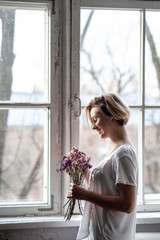Smiling young woman near the window holds a bouquet of purple flowers near her face  - 324882194