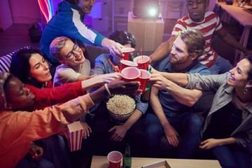 Group of young people toasting with beer while sitting at home with popcorn and they are going to watch a movie