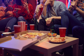 Close-up of pizza and drinks are on the table with young people sitting on sofa eating and drinking...