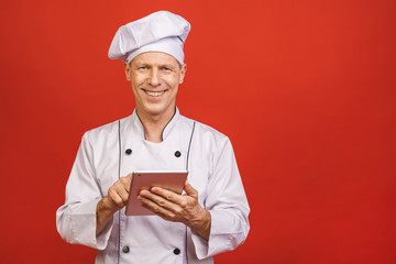 Portrait of a smiling senior smiling chef cook holding tablet computer isolated on a red background.