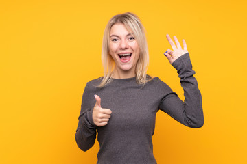 Young blonde woman over isolated yellow background showing ok sign and thumb up gesture