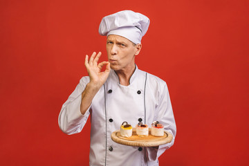A portrait of a senior baker holding delicious cakes isolated on red background.