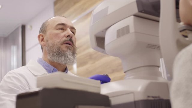 Low angle shot of senior male optometrist in lab coat using autorefractor while checking eyes of little patient in ophthalmology clinic