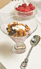Top, front view of a clear, glasscup filled with frreshly baked, homemade, cerialMaple Quinoa Granola,with arn artisan spoon on a white table cloth