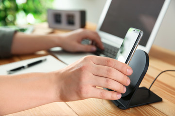 Man putting mobile phone onto wireless charger at wooden table, closeup. Modern workplace accessory