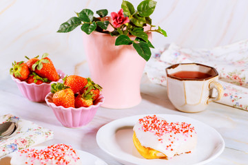 Two sweet heart shaped donut with white glaze and sprinkles on party table with tea, strawberries and rose.