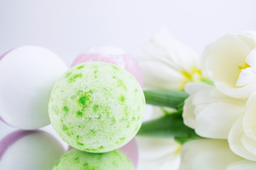 Obraz na płótnie Canvas A green and pink salt bomb on a white gray background is reflected, next to white tulips. Mineral makeup. Banner. Spa care. Banner. Copy space