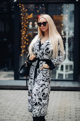 Stock photo of a gorgeous young adult woman with long straight blonde hair wearing black sunglasses, black and white coat with belt and fur cuffs and carrying a black classic handbag. She is standing