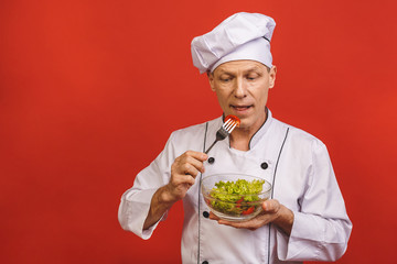 Picture of happy young senior chief cook in uniform standing isolated over red wall background, holding and eating salad.