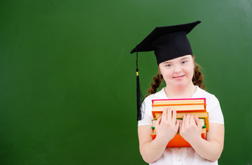 A girl with down syndrome stands against a blackboard with books in a graduate’s hat. Smiling while looking at the camera.