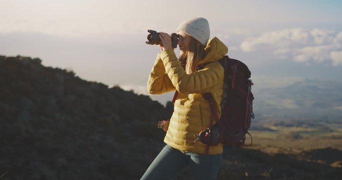 Young adventurous woman in a yellow jacket hiking in the mountains at sunset taking photographs with a digital camera, creative photographer taking photos in the great outdoors