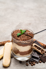 Classic tiramisu dessert in a glass cup, pieces of chocolate and savoiardi cookies on concrete background