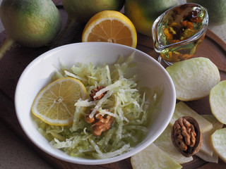 Salad of raw grated green radish, walnuts, lemon in a white bowl, vegetables, radish slices, oil on a wooden stand closeup. Recipe for a vegetarian dish for proper nutrition.