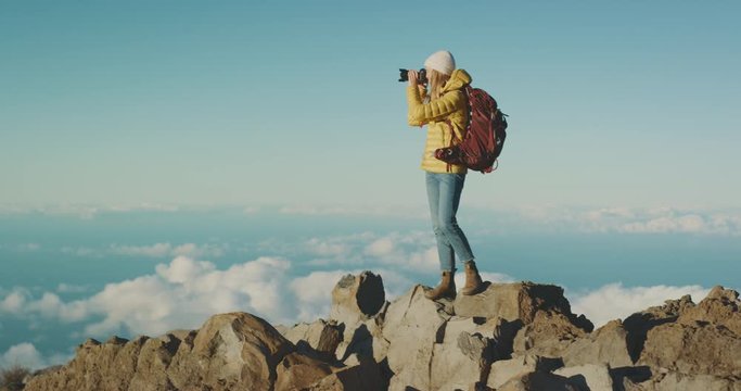 Young adventurous photographer woman standing on a mountain top above the clouds taking photos, backpacking traveler exploring the outdoors