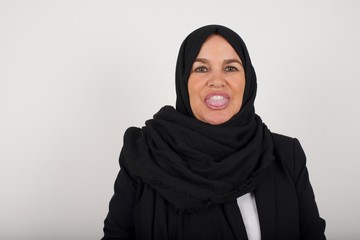 Beautiful muslim woman with happy and funny face smiling and showing tongue. Wearing casual clothes and standing against gray studio background.