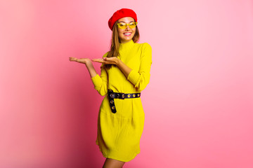 Beautiful young woman in acid yellow mini dress and red hat is looking at camera, pointing and smiling. Three quarter length studio shot on pink background.