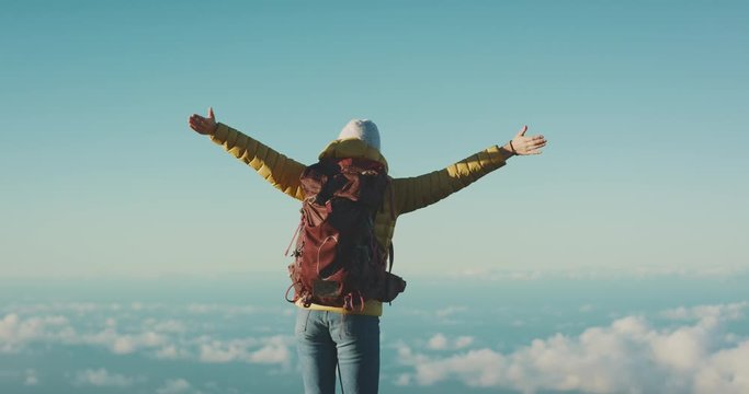 Young woman wearing a yellow jacket and hiking backpack raising her arms into the sky and enjoying the view above the clouds on top of a mountain