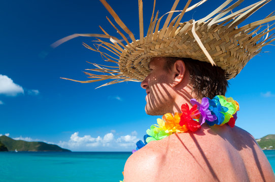 Tourist standing in front of a vibrant blue tropical paradise wearing woven palm frond hat and gaycation rainbow flower lei