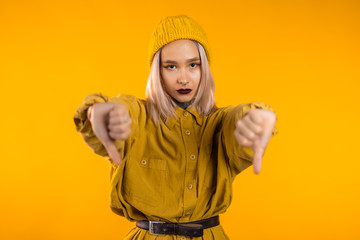 Young pretty woman standing on yellow studio background expressing discontent and showing thumb down gesture at camera. Portrait of girl with sign of dislike
