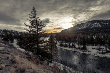 Early morning sunrise over river running through tree covered valley with snow capped mountains and clouds