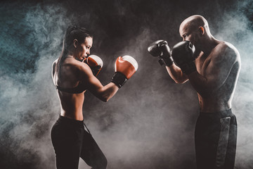 Fototapeta na wymiar Shirtless Woman exercising with trainer at boxing and self defense lesson, studio, smoke on background. Female and male fight,