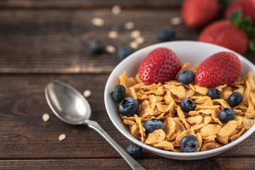 Granola chips with blueberries and raspberries in white bowl in dark wooden desk.