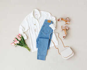 Blue jeans, white shirt, heeled sandals, bag with chain strap, jewelry, bouquet of pink tulips flowers on beige background. Women's stylish spring summer outfit. Trendy clothes. Flat lay, top view.