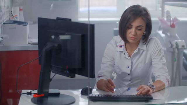 Professional female doctor in a lab coat, typing on a computer with a monitor sitting behind a glass wall in the office.