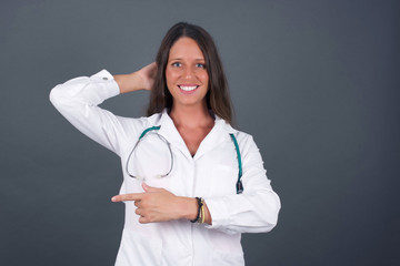 Joyful pretty young doctor woman wearing medical uniform demonstrates something. One hand on her head and pointing with other hand.