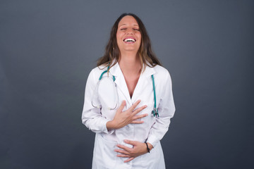 Joyful European doctor woman keeps hands crossed, laughs at good joke, wears casual clothes and round spectacles, standing indoors. Happy young woman with long hair poses against gray wall.